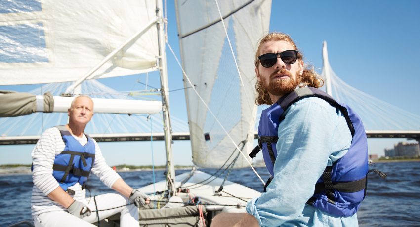 Serious handsome men in life jacket frowning from sun looking around while traveling by sail boat on river